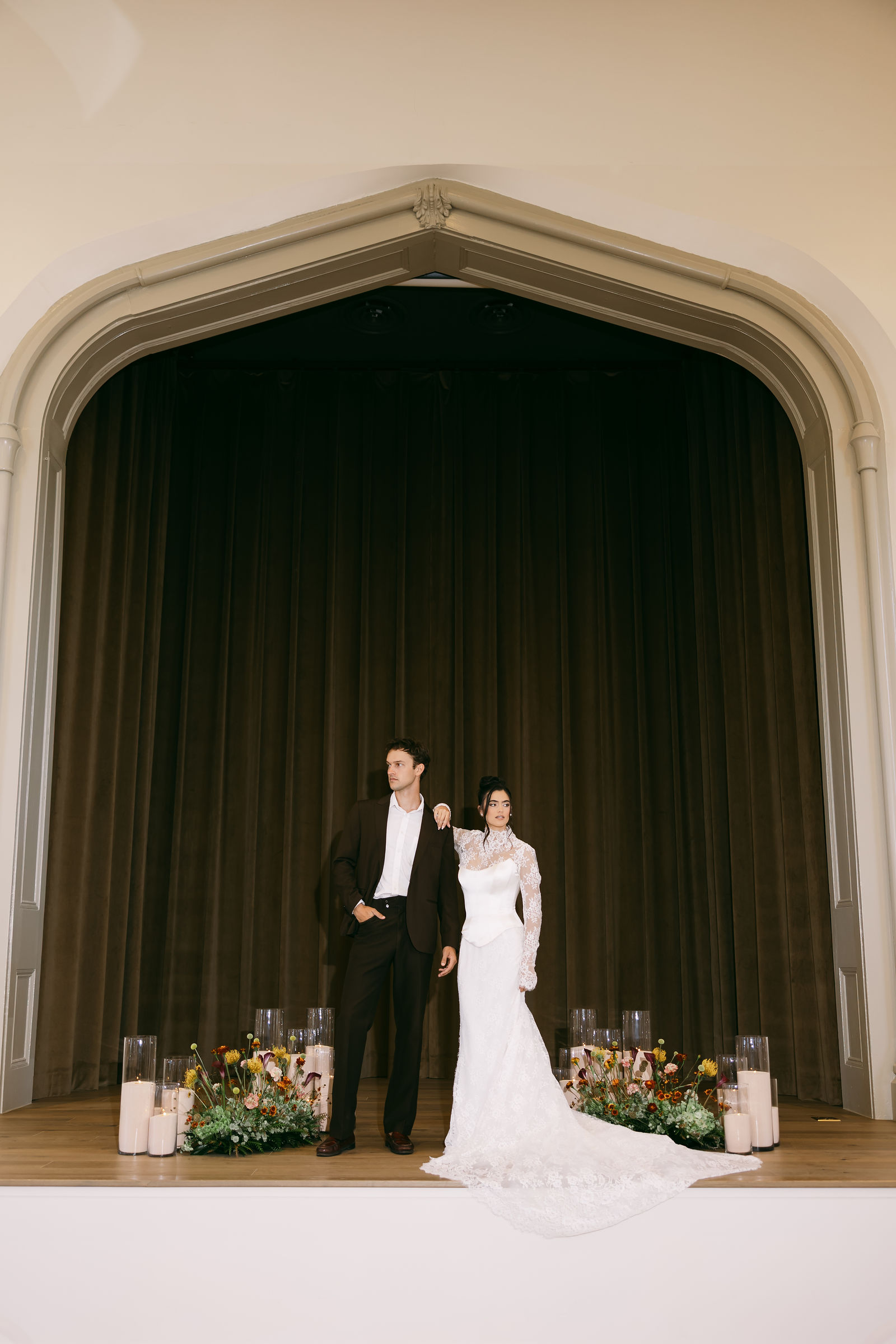 Bride and groom standing together on a candlelit stage beneath an ornate arch, framed by autumn floral arrangements